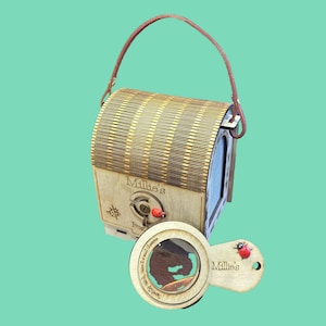 May include: A wooden bug observation box with a handle and a magnifying glass. The box is made of light-colored wood with a slatted top and the name "Millie's" is engraved on the front. A small ladybug is attached to the front. The magnifying glass also has the name "Millie's".