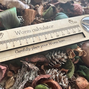 Puede incluir: Un calculador de gusanos de madera con una lupa y una regla marcada en centímetros. El texto en la regla dice "Worm calculator" y "Carefully place your Worm here..."