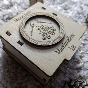 May include: A wooden box with a personalised name, a campfire design, and the text "Marshmallow kit".