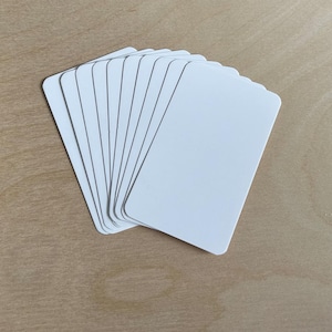 Puede incluir: Una pila de tarjetas blancas en blanco con esquinas redondeadas, ligeramente abanicadas. Las tarjetas están dispuestas sobre una superficie de madera de color marrón claro. Las tarjetas son de color blanco brillante.