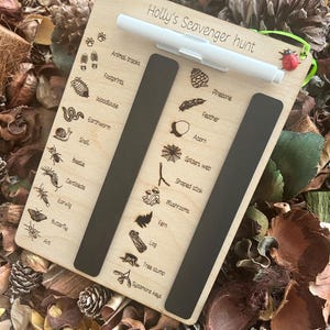 A wooden scavenger hunt board with various animal and plant illustrations, placed on a bed of pine needles and leaves.