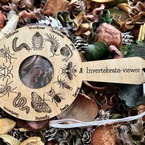 Könnte beinhalten: Eine Lupe aus Holz mit einer klaren Linse und einem Griff, auf dem "Invertebrate-viewer" steht. Der Griff ist mit einem Kreis aus verschiedenen Wirbellosen verziert, darunter eine Biene, ein Schmetterling, ein Käfer, eine Spinne, ein Tausendfüßler, ein Assel, eine Wanze, eine Ohrwurm, eine Schnecke und eine Ameise.