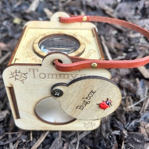 May include: A wooden bug box with a magnifying glass, a red leather strap, and the name "Tommy" engraved on the side. The box features a small ladybug illustration and the words "Bug box".