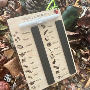 A wooden scavenger hunt board with various animal and plant illustrations, placed on a bed of pine needles and leaves.