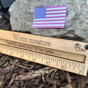 May include: Wooden worm measure with engraved text and ruler markings. The text reads "Worm measure" and "Carefully place your Worm here...". An American flag sticker is in the background. The ruler is approximately 7 inches long.