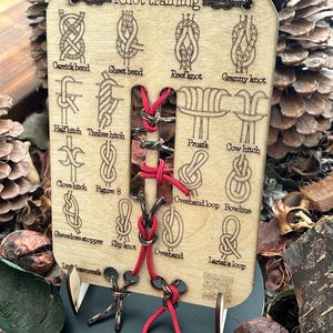 Puede incluir: Tablero de entrenamiento de nudos de madera con cuerda roja y negra, que muestra diferentes nudos con sus nombres, incluyendo Carrick bend, Sheet bend, Reef knot, Granny knot, Half hitch, Timber hitch, Prusik, Cow hitch, Clove hitch, Figure 8, Overhand loop, Bowline, Stevedore stopper, Slip knot, Overhand, y Lariat's loop.