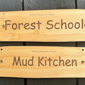 May include: Two rectangular light-coloured wooden signs with irregular, jagged edges. The top sign is engraved with "Tarleton Community Primary" and "Forest School". The bottom sign is engraved with "Tarleton Community Primary" and "Mud Kitchen".