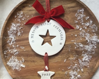 May include: A white ceramic Christmas ornament with a star cutout and the text "Heaven's First Christmas". It is adorned with a red velvet bow and a small star pendant with "2022" inscribed. The ornament hangs from a red ribbon and rests on a wooden plate.