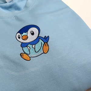Puede incluir: Sudadera azul claro con un bordado de un personaje de dibujos animados de pingüino. El pingüino tiene una cara blanca, detalles azules y negros y pies naranjas. La sudadera es de material suave y tiene cuello redondo.
