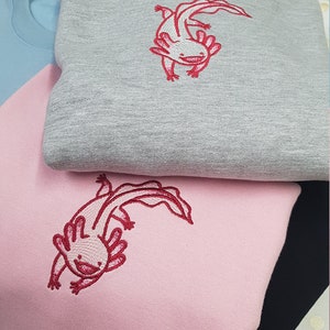 Puede incluir: Tres sudaderas con bordado de ajolote rosa. Las sudaderas son grises, rosas y negras.