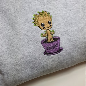 May include: A gray sweatshirt with an embroidered image of a small, brown, cartoon character with green hair and a purple pot. The pot has the word "GROOT" written on it.
