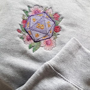 Puede incluir: Una sudadera gris claro con un dado de veinte caras bordado en morado y dorado rodeado de flores rosas y blancas.