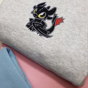Puede incluir: Sudadera gris claro con un bordado de un dragón negro con ojos amarillos y detalles rojos. La sudadera está doblada sobre una sudadera rosa y azul.