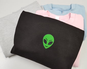 May include: A stack of sweatshirts in various colors, including black, gray, light blue, and pink. The black sweatshirt has a green alien face embroidered on the front.