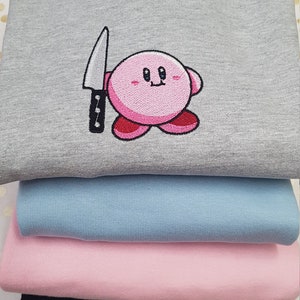 Puede incluir: Una sudadera gris con un personaje de dibujos animados rosa que sostiene un cuchillo bordado en la parte delantera. La sudadera está apilada encima de una sudadera azul claro, una sudadera rosa y una sudadera negra.