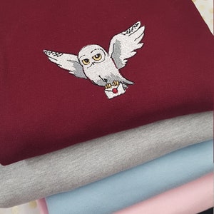 Puede incluir: Una pila de camisetas de varios colores, incluyendo burdeos, gris, azul claro, rosa y negro. La camiseta de arriba tiene un búho blanco bordado que lleva un sobre rojo.