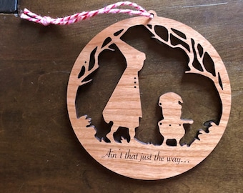 Puede incluir: Adorno redondo de madera con un diseño recortado que representa dos figuras bajo un árbol. El adorno tiene una cuerda roja y blanca para colgar y el texto "Ain't that just the way..." está grabado en la madera.