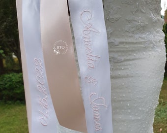 May include: Three white satin ribbons with pink embroidery. The ribbons are tied together and have the names "Olivia & James" and "Alexa" written on them.