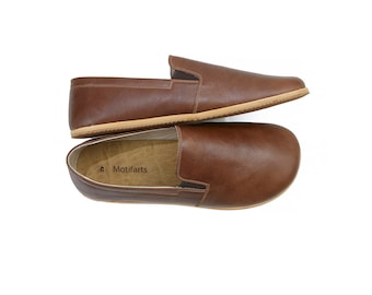 May include: A pair of brown leather slip-on shoes with a light brown sole. The shoes are laid out on a white background. The brand name "Motifarts" is printed on the insole of one shoe.