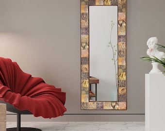 Puede incluir: Un espejo alto y rectangular con un marco decorativo hecho de pequeños azulejos con patrones intrincados en tonos marrones, dorados y crema. El espejo refleja una habitación con una silla roja y una planta.