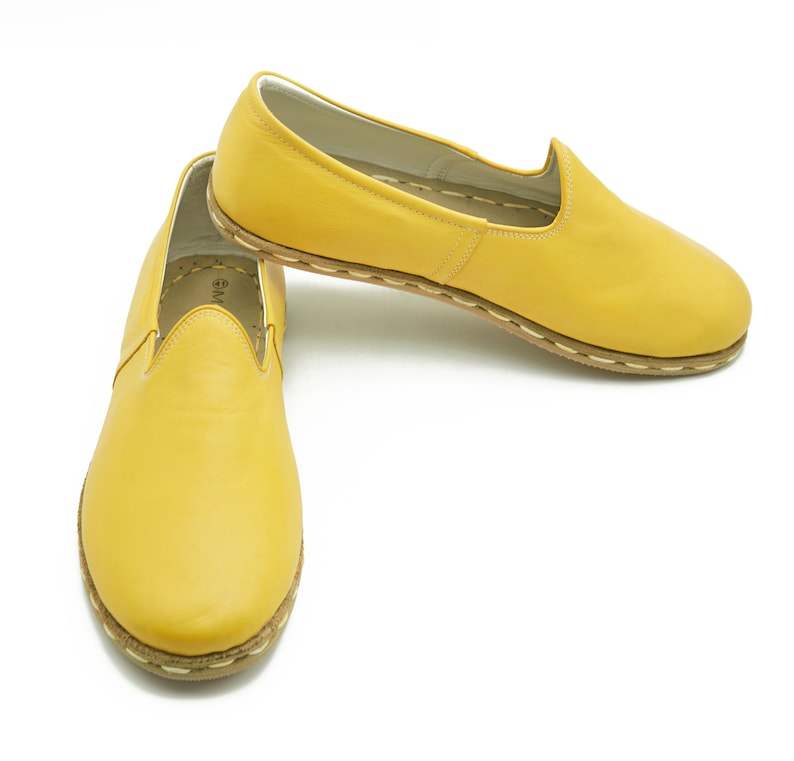 Puede incluir: Un par de zapatos amarillos de cuero sin cordones con un detalle de puntadas blancas alrededor de la suela.