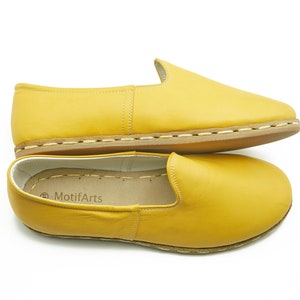 Puede incluir: Un par de zapatos amarillos de cuero con suela cosida marr&oacute;n. Los zapatos est&aacute;n apilados uno encima del otro y el nombre de la marca "MotifArts" es visible en la plantilla del zapato inferior.