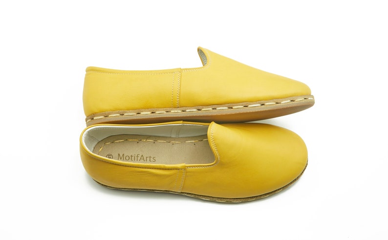 Puede incluir: Un par de zapatos amarillos de cuero con suela cosida marr&oacute;n. Los zapatos est&aacute;n apilados uno encima del otro y el nombre de la marca "MotifArts" es visible en la plantilla del zapato inferior.