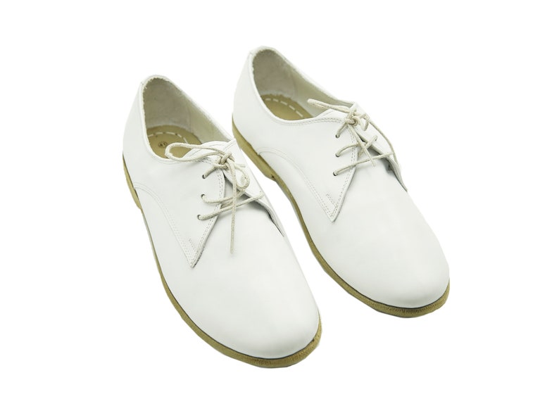 Puede incluir: Un par de zapatos blancos de cuero con cordones y suela de color crema.