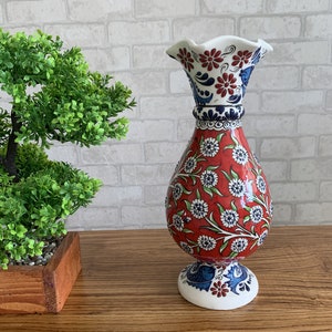 Könnte beinhalten: Eine rote und weiße Keramikvase mit einem Blumenmuster. Die Vase hat eine breite Öffnung und einen schmalen Boden. Sie ist mit blauen und grünen Akzenten verziert.