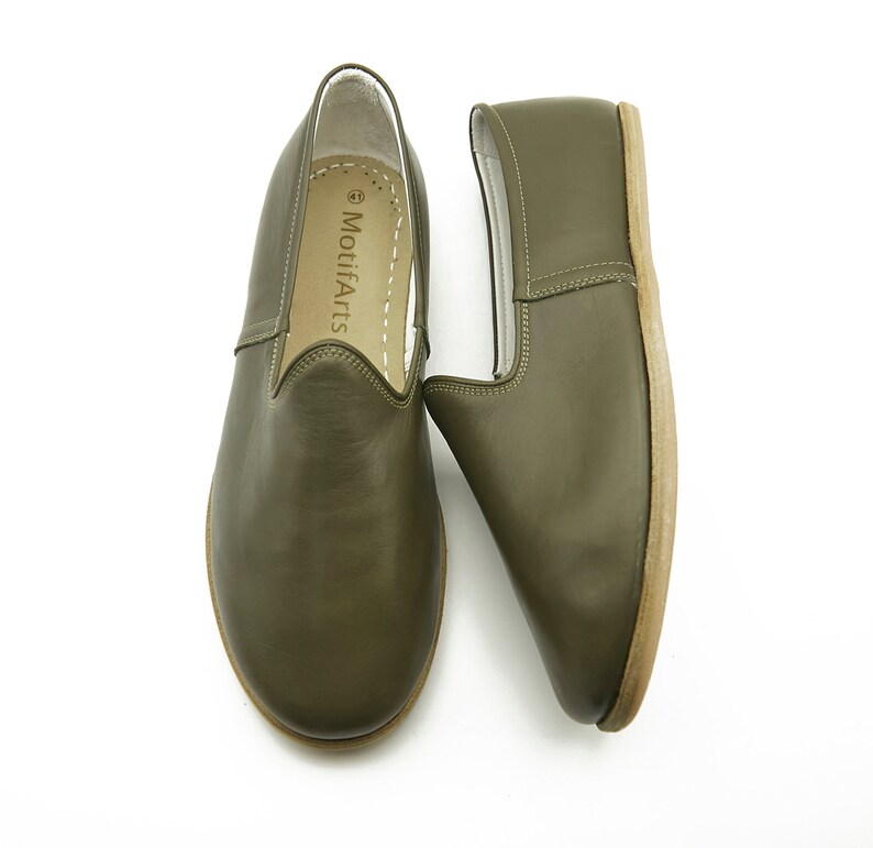 K&ouml;nnte beinhalten: Ein Paar olivgr&uuml;ne Slipper aus Leder mit wei&szlig;em Lederfutter. Die Schuhe haben ein schlichtes Design und sind aus weichem, geschmeidigem Leder gefertigt. Der Markenname "MotifArts" ist auf der Innenseite des Schuhs aufgedruckt.