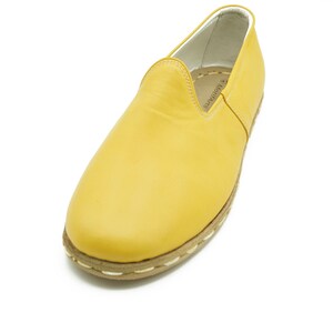 Puede incluir: Un par de zapatos de cuero amarillo de vestir sin cordones con un ribete blanco cosido alrededor de la suela. Los zapatos tienen un dise&ntilde;o sencillo y est&aacute;n hechos de cuero suave.