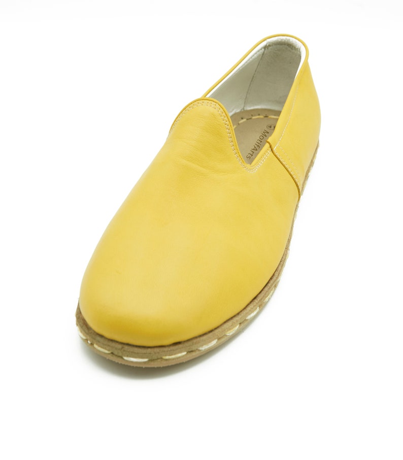 Puede incluir: Un par de zapatos de cuero amarillo de vestir sin cordones con un ribete blanco cosido alrededor de la suela. Los zapatos tienen un dise&ntilde;o sencillo y est&aacute;n hechos de cuero suave.