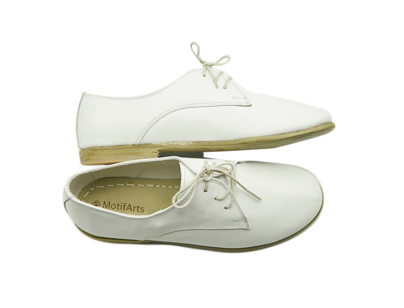 Puede incluir: Un par de zapatos blancos de cuero con cordones y suela marr&oacute;n. Los zapatos est&aacute;n apilados uno encima del otro. La marca "MotifArts" es visible en el interior del zapato.