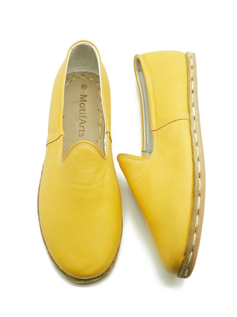 Puede incluir: Un par de zapatos de cuero amarillo de vestir sin cordones con detalles de costura blanca alrededor de la suela. Los zapatos tienen un dise&ntilde;o sencillo y est&aacute;n hechos de cuero suave. El nombre de la marca "MotifArts" est&aacute; impreso en el interior del zapato.