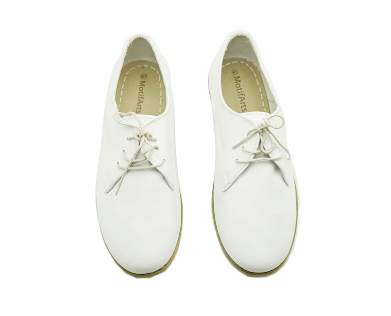 Puede incluir: Un par de zapatos de cuero blanco con cordones y suela marr&oacute;n. Los zapatos tienen el texto "MotifArts" impreso en el interior de cada zapato.