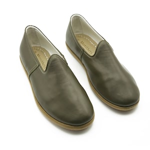 K&ouml;nnte beinhalten: Ein Paar olivgr&uuml;ne Lederschlappen mit brauner Sohle. Die Schuhe haben den Text "MoufArts" auf der Innenseite jedes Schuhs aufgedruckt.