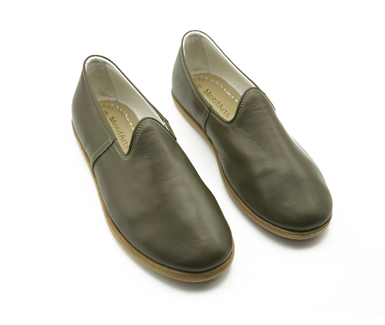 K&ouml;nnte beinhalten: Ein Paar olivgr&uuml;ne Lederschlappen mit brauner Sohle. Die Schuhe haben den Text "MoufArts" auf der Innenseite jedes Schuhs aufgedruckt.