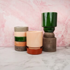 Könnte beinhalten: Ein Set aus vier Keramik-Blumentöpfen in verschiedenen Farben, darunter Grün, Braun, Beige und Orange. Die Töpfe sind übereinander gestapelt, wobei der größte Topf unten und der kleinste Topf oben ist. Die Töpfe sind alle rund und haben eine glatte Oberfläche.