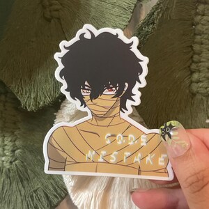 Puede incluir: Una pegatina con un personaje de dibujos animados con cabello negro, ojos rojos y vendas envueltas alrededor de su cabeza y torso. El personaje lleva una camisa blanca con el texto "GOD'S MISTAKE" impreso en ella.