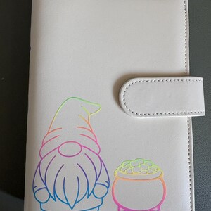 May include: A light grey rectangular notebook with a magnetic strap closure. The front cover features a colourful gradient outline graphic of a gnome standing next to a small pot filled with round objects, rendered in rainbow-coloured lines.
