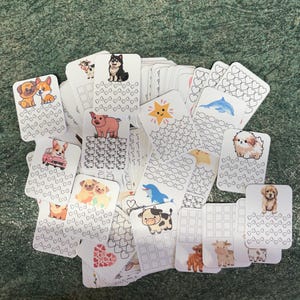 May include: A pile of white, rectangular cards featuring animal illustrations, including dogs, pigs, and cows. The cards display colourful drawings and decorative patterns, such as hearts, stars, and bow ties. Some cards have grid patterns.