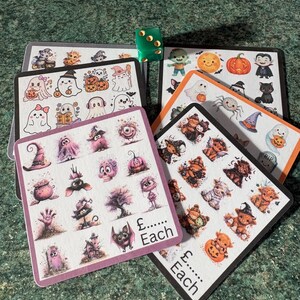 May include: A collection of square sticker sheets featuring various Halloween-themed illustrations including ghosts, pumpkins, bats, skulls, and creatures. Each sheet displays the text £...... Each. A single green six-sided die rests on top of the cards.