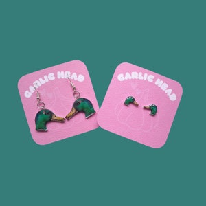Op de afbeelding: Een paar hangende oorbellen met een groen en blauw eendenkopontwerp. De oorbellen bevinden zich op roze kaartjes met de tekst "Garlic Head" erop gedrukt.
