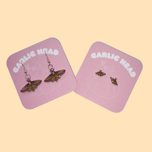 Puede incluir: Dos pares de pendientes con un diseño de polilla. Los pendientes están en tarjetas rosas con el texto "Garlic Head" impreso en ellas.