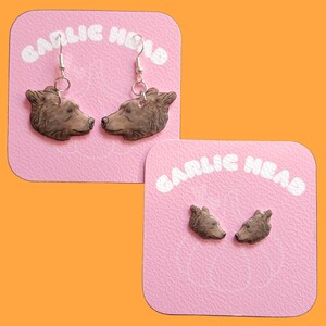 Puede incluir: Pendientes y pendientes de cabeza de oso marrón exhibidos en tarjetas rosas con el texto "GARLIC HEAD" en blanco. Los pendientes están sobre un fondo naranja.