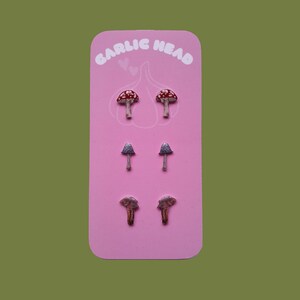 May include: A pink card with the text "GARLIC HEAD" in white letters. The card features three pairs of mushroom-shaped earrings. The first pair is red with white spots, the second pair is grey, and the third pair is brown.