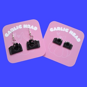 Puede incluir: Dos pares de pendientes de cámara negra en tarjetas rosas con el texto "Garlic Head" impreso en ellas.