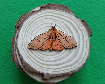 Puede incluir: Un broche de madera con un diseño estilizado de polilla. La polilla tiene detalles en naranja y negro, con un contorno negro. El broche está colocado sobre una pequeña rodaja de madera redonda con anillos de crecimiento visibles. El fondo es de color verde liso.