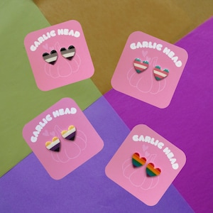 Könnte beinhalten: Vier rosa Karten mit dem aufgedruckten Text "Garlic Head". Jede Karte zeigt ein Paar herzförmiger Ohrringe mit verschiedenen Pride-Flaggen-Designs: Schwarz, Weiß und Lila; Grün, Weiß und Rosa; Gelb, Weiß und Lila; und Regenbogen.