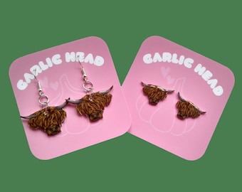 May include: A pair of dangle earrings featuring a Highland Cow head design. The earrings are silver with a pink background and the text "GARLIC HEAD" is printed on the background.